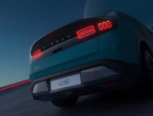 Nahaufnahme des Hecks eines blauen Nissan Leaf-Elektroautos in der Dämmerung, mit beleuchteten Rückleuchten und einem deutlich sichtbaren LEAF-Schriftzug. Der Himmel ist dunkel mit einem großen Mond im Hintergrund.