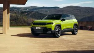 Ein leuchtend grüner Jeep-Geländewagen parkt auf einer großen Steinterrasse mit malerischen, baumbewachsenen Hügeln und einem teilweise bewölkten Himmel im Hintergrund. Das Fahrzeug ist modern mit schwarzen Akzenten und großen Rädern.