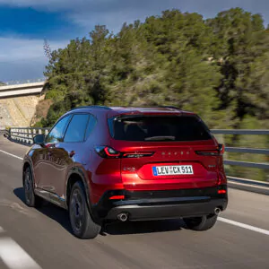 Ein roter Mazda CX-5 SUV, der im Rahmen des Mazda CX-5-Leasings erhältlich ist, fährt auf einer Autobahn neben einer Leitplanke, im Hintergrund sind Bäume und eine kleine Brücke vor blauem Himmel zu sehen.