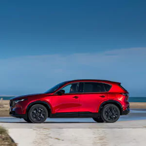 Ein leuchtend roter SUV, der im Rahmen des Mazda CX-5-Leasings erhältlich ist, fährt auf einer asphaltierten Straße in Strandnähe, im Hintergrund sind das Meer und ein strahlend blauer Himmel zu sehen.