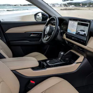Der Innenraum eines modernen Mazda CX-5-Leasingfahrzeugs verfügt über beigefarbene Sitze, ein digitales Armaturenbrett sowie ein großes Touchscreen-Display, auf dem eine Karte und eine Fahrzeugansicht angezeigt werden, und bietet einen atemberaubenden Blick auf den Strand und das Meer vor dem Fenster.