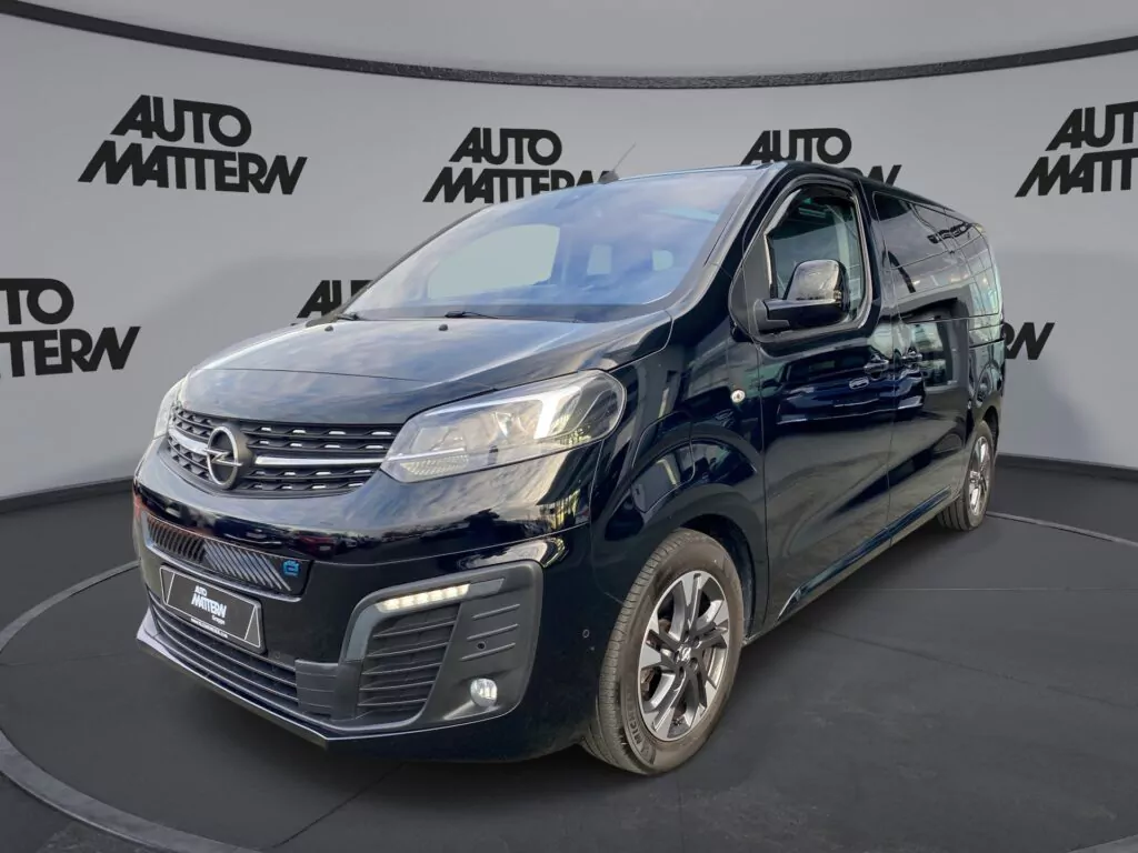 Ein schwarzer Opel Transporter parkt in einem überdachten Ausstellungsraum mit Auto Mattern-Logos an den Wänden im Hintergrund, aufgenommen aus dem Blickwinkel von vorne links. Besuchen Sie uns am Tag der offenen Tür März 2026, um ihn aus der Nähe zu sehen!.