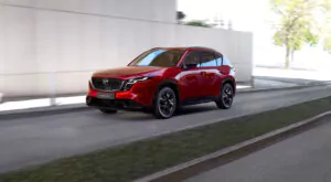 Ein roter Mazda CX-5 SUV fährt auf einer Stadtstraße neben einer Betonmauer, die Unschärfe der Umgebung deutet auf Bewegung hin - verpassen Sie nicht unseren Tag der offenen Tür März 2026, um diese dynamische Performance live zu erleben.