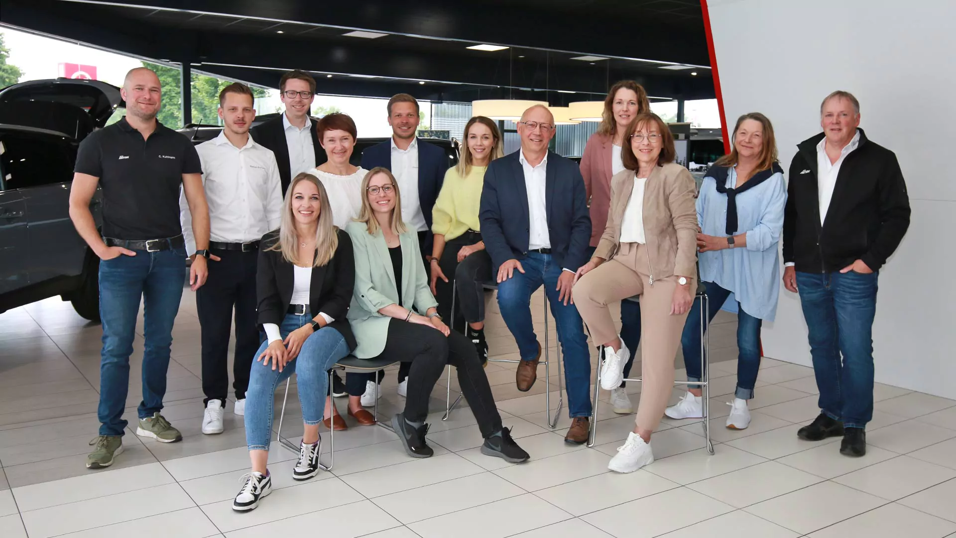 Eine Gruppe von dreizehn Personen posiert und lächelt zusammen in einem modernen Ausstellungsraum mit Autos im Hintergrund; die meisten stehen, während einige auf Hockern sitzen. Die Umgebung ist hell und professionell.