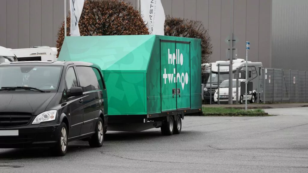 Ein schwarzer Lieferwagen zieht einen großen grünen Anhänger mit der Aufschrift "Hello Twike", der auf einer grauen Straße neben Wohnmobilen und Industriegebäuden im Hintergrund geparkt ist.