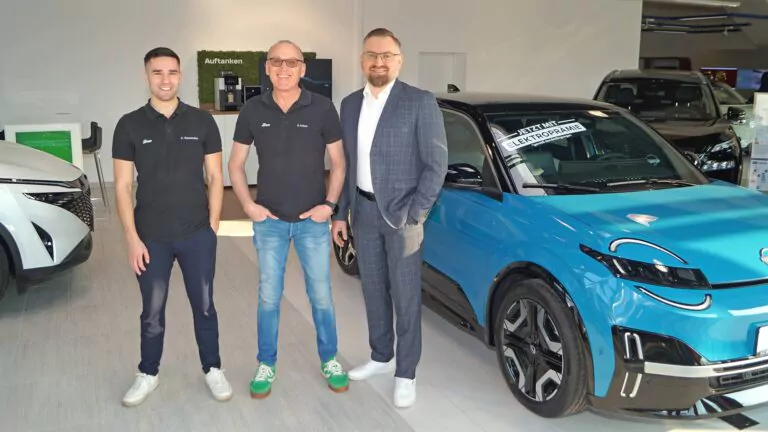 Drei Männer stehen lächelnd in einem modernen Autohaus neben einem blauen Elektroauto. Zwei Männer tragen schwarze Polohemden und Jeans, der dritte trägt einen Anzug. Im Hintergrund sind weitere Autos und Ladestationen für Elektroautos zu sehen.