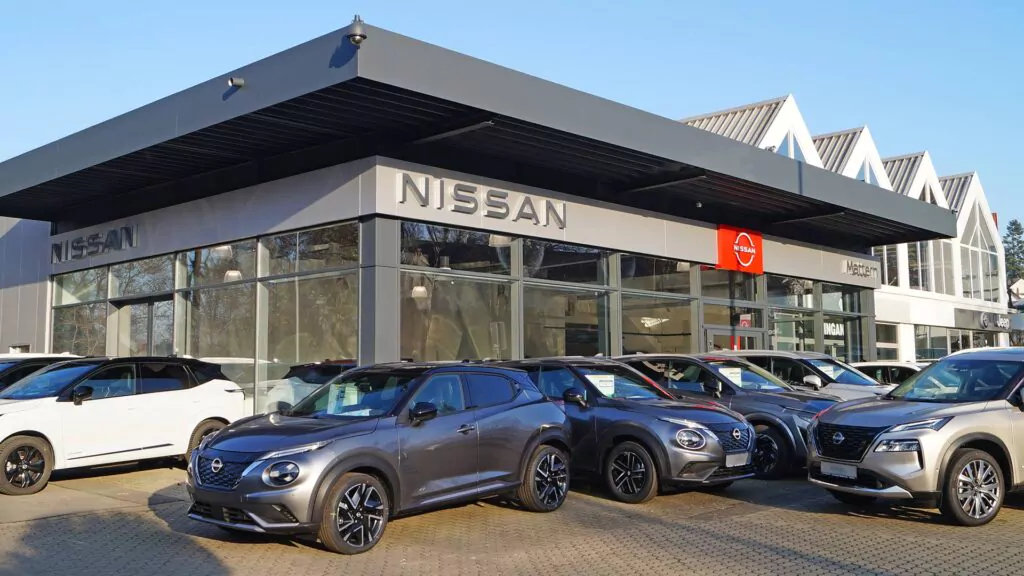 Mehrere neue Nissan-Fahrzeuge sind vor einem modernen Nissan-Händlergebäude mit großen Fenstern und einem auffälligen Nissan-Logo an der Fassade geparkt.