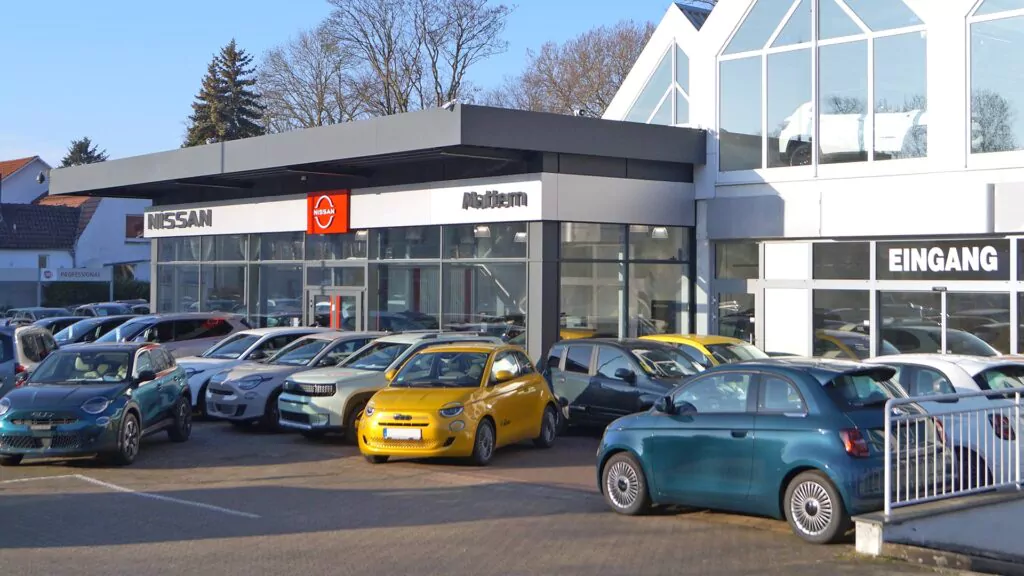 Ein Autohaus mit mehreren geparkten Autos davor, darunter ein leuchtend gelbes Auto. Auf dem Gebäude sind die Logos von Nissan und AutoTeam zu sehen, und ein Schild mit der Aufschrift "Eingang" ist sichtbar. Der Bereich ist gut beleuchtet und modern.