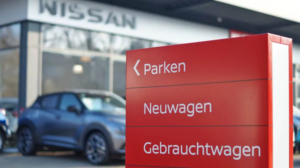Ein rotes Schild an einem Nissan-Händler zeigt eine Wegbeschreibung in deutscher Sprache an: Parken, Neuwagen und Gebrauchtwagen, mit einem unscharfen Auto und dem Gebäude des Händlers im Hintergrund.