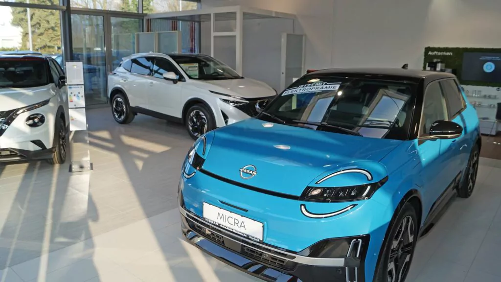 Ein blauer Nissan Micra wird in einem hellen Autohaus neben einem weißen Geländewagen und einem weiteren weißen Auto ausgestellt, dessen große Fenster viel Sonnenlicht hereinlassen. Im Inneren des Ausstellungsraums sind Schilder und Informationsdisplays zu sehen.