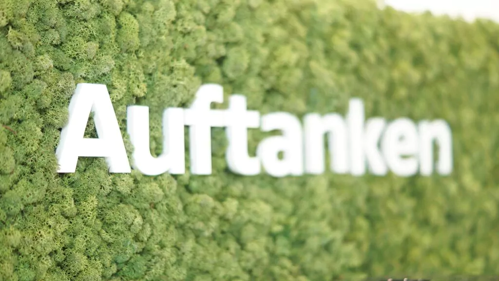 Weiße 3D-Buchstaben mit der Aufschrift Auftanken sind auf einer grünen, strukturierten Wand montiert, die mit Moos oder künstlichen Pflanzen bedeckt ist. Das Wort ist scharf, während der Hintergrund leicht unscharf ist.