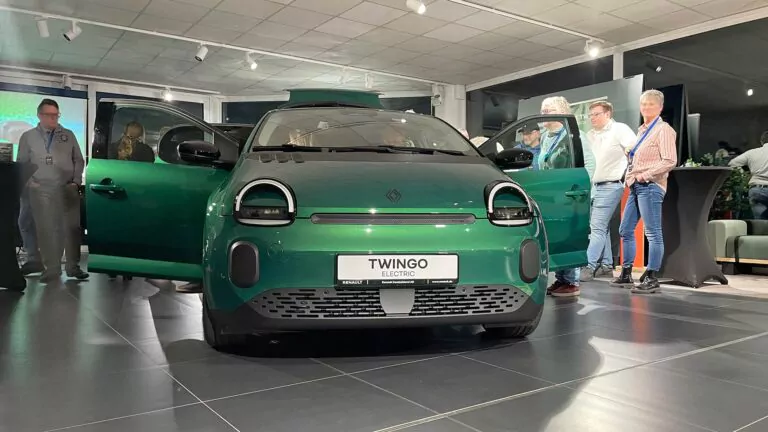 Ein grüner Renault Twingo Electric ist in einem Ausstellungsraum ausgestellt, die Türen sind geöffnet. Mehrere Personen stehen um das Auto herum, beobachten es und unterhalten sich in einem gut beleuchteten Ausstellungsraum.