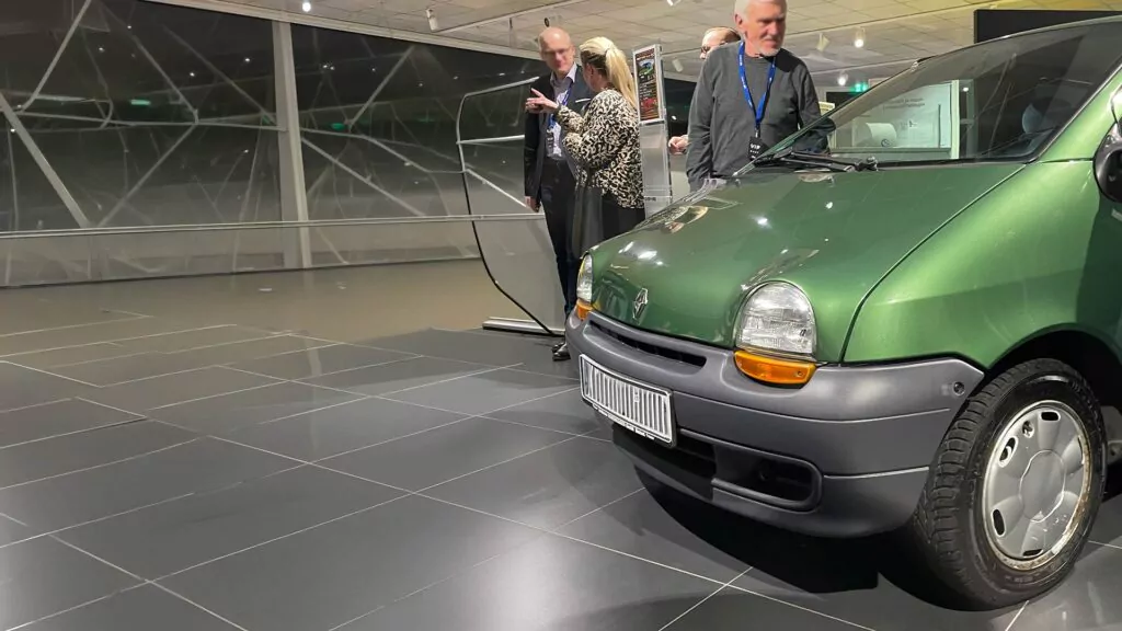 Eine Gruppe von drei Personen steht und unterhält sich neben einer Auslage in einem modernen Ausstellungsraum; im Vordergrund ist die Front eines grünen Autos auf glänzenden schwarzen Fliesen zu sehen.