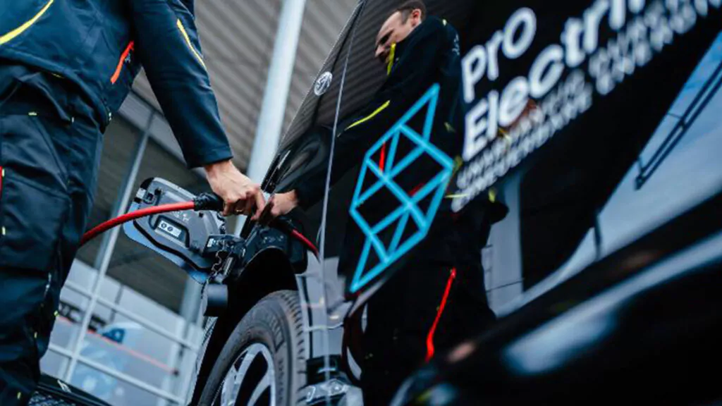 Eine Person lädt ein Elektrofahrzeug an einer Ladestation auf. Das Auto trägt den Schriftzug Pro Electric und geometrische Motive an der Seite. Die Szene spielt in einem modernen, überdachten Bereich.