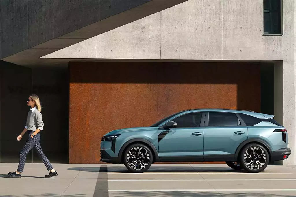 Eine Frau geht an einem geparkten hellblauen Dacia Striker mit schwarzen Rädern vorbei, der vor einem modernen Gebäude aus Beton und rostfarbenen Wänden steht.