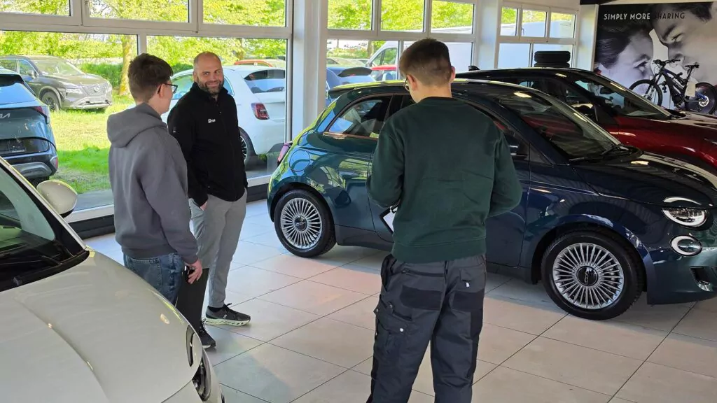 Drei Männer unterhalten sich in einem Autohaus. Sie stehen in der Nähe eines dunkelblauen Kleinwagens, in dessen Innenraum und durch große Fenster weitere Fahrzeuge zu sehen sind. Helles Tageslicht strömt herein und beleuchtet den Ausstellungsraum.