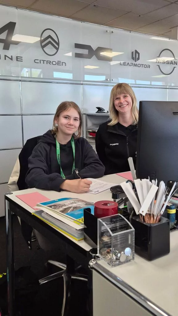 Zwei Frauen sitzen an einem modernen Bürotisch, eine schreibt in ein Notizbuch, die andere lächelt in die Kamera. Auf dem Schreibtisch liegen Büromaterial und Ordner, an der Glaswand hinter ihnen sind Logos von Autofirmen zu sehen.