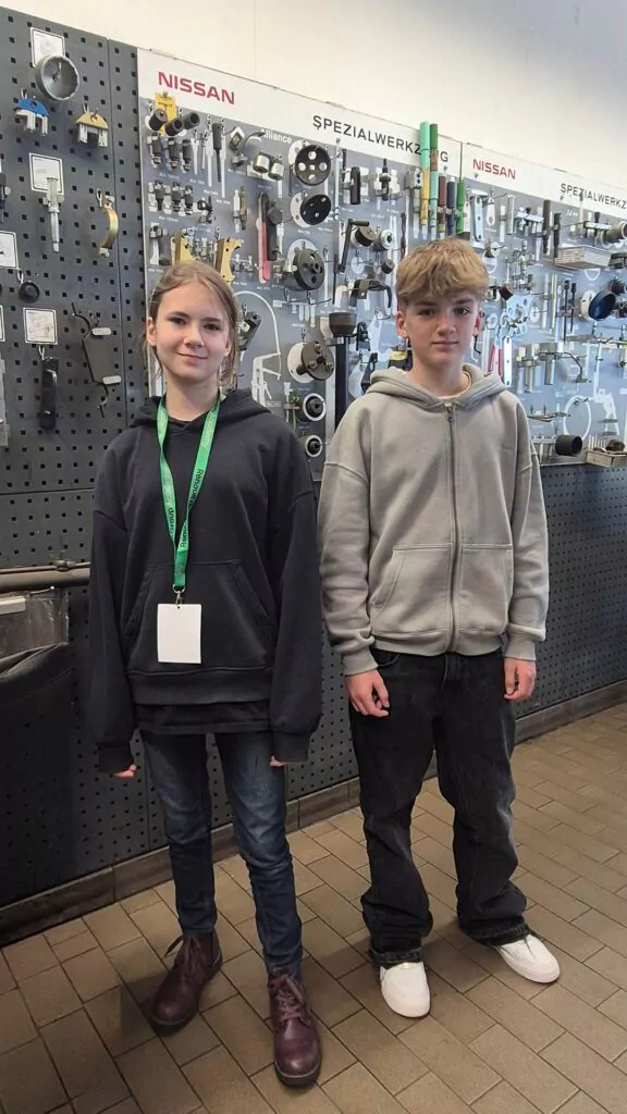 Zwei Teenager stehen nebeneinander vor einer Wand, die mit verschiedenen Werkzeugen und Autoteilen bedeckt ist. Beide tragen Kapuzenpullis und Jeans; einer hat ein grünes Schlüsselband und ein Abzeichen, der andere hat die Hände in den Taschen.