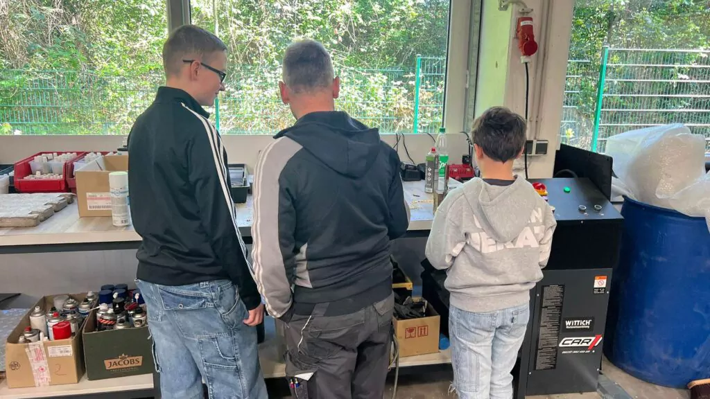 Drei Personen, von hinten gesehen, stehen an einer Werkbank in einer Werkstatt oder einem Labor am Girls & Boys Day. Auf der Theke stehen verschiedene Flaschen und Geräte. Große Fenster geben den Blick auf Grünpflanzen und einen Drahtzaun frei.