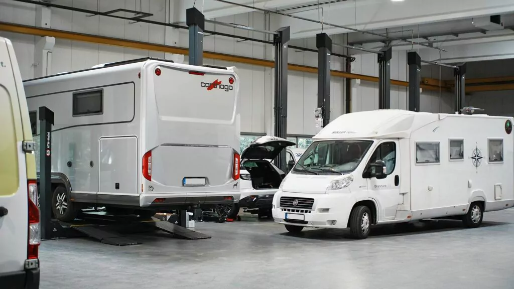 Zwei weiße Wohnmobile stehen in einer gut beleuchteten Garage. Ein Fahrzeug steht auf einer Hebebühne, während das andere auf dem Boden geparkt ist. Im Hintergrund sind verschiedene Kfz-Geräte und Werkzeuge zu sehen.