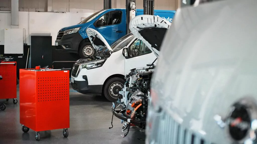 In einer Werkstatt werden mehrere Lieferwagen mit offenen Motorhauben gewartet. Zwei rote Werkzeugschränke stehen in der Nähe, und die Werkstatt macht einen sauberen, organisierten Eindruck. Der Schwerpunkt liegt auf der Wartung und Reparatur von Fahrzeugen.