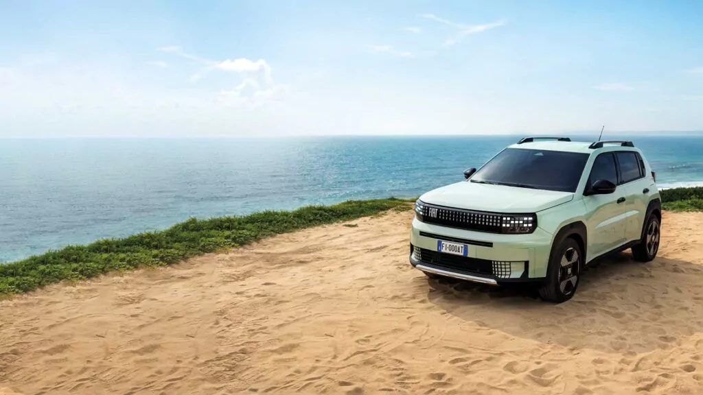 Ein hellgrüner Geländewagen parkt auf einer sandigen Fläche in der Nähe eines Grünstreifens und überblickt das ruhige blaue Meer unter einem meist klaren Himmel.