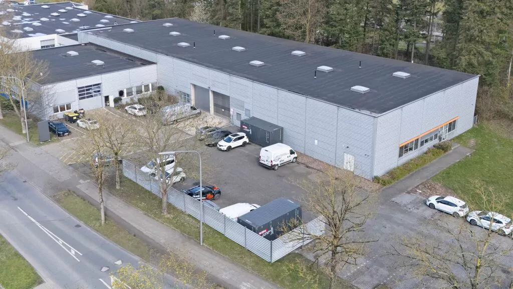 Eine große Industriehalle mit schwarzem Flachdach und grauer Metallverkleidung, umgeben von einigen geparkten Autos und Bäumen. Das Gebäude beherbergt eine Nutzfahrzeug Wohnmobil Werkstatt, grenzt an eine Straße und ist in einigen Bereichen von einem Zaun umgeben.