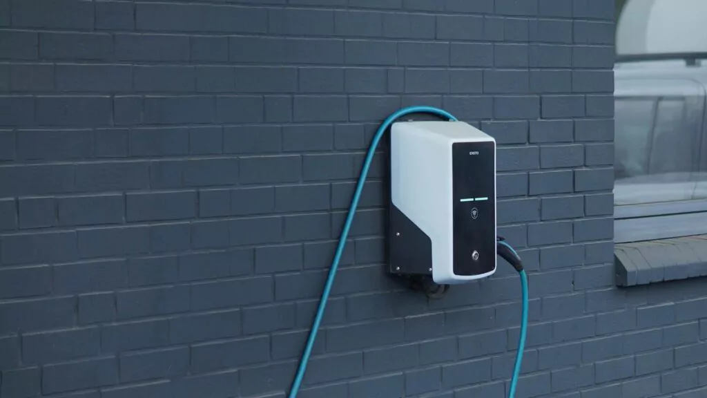 Eine weiß-schwarze Ladestation für Elektrofahrzeuge mit blauem Kabel, ideal für die Wallbox Förderung E-Auto, ist an einer dunkelgrauen Backsteinwand neben einem Fenster angebracht.