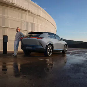 Eine hell gekleidete Person lehnt sich an einen silbernen Elektro-Geländewagen, der auf einer nassen Fläche in der Nähe eines modernen, runden Gebäudes unter freiem Himmel geparkt ist.