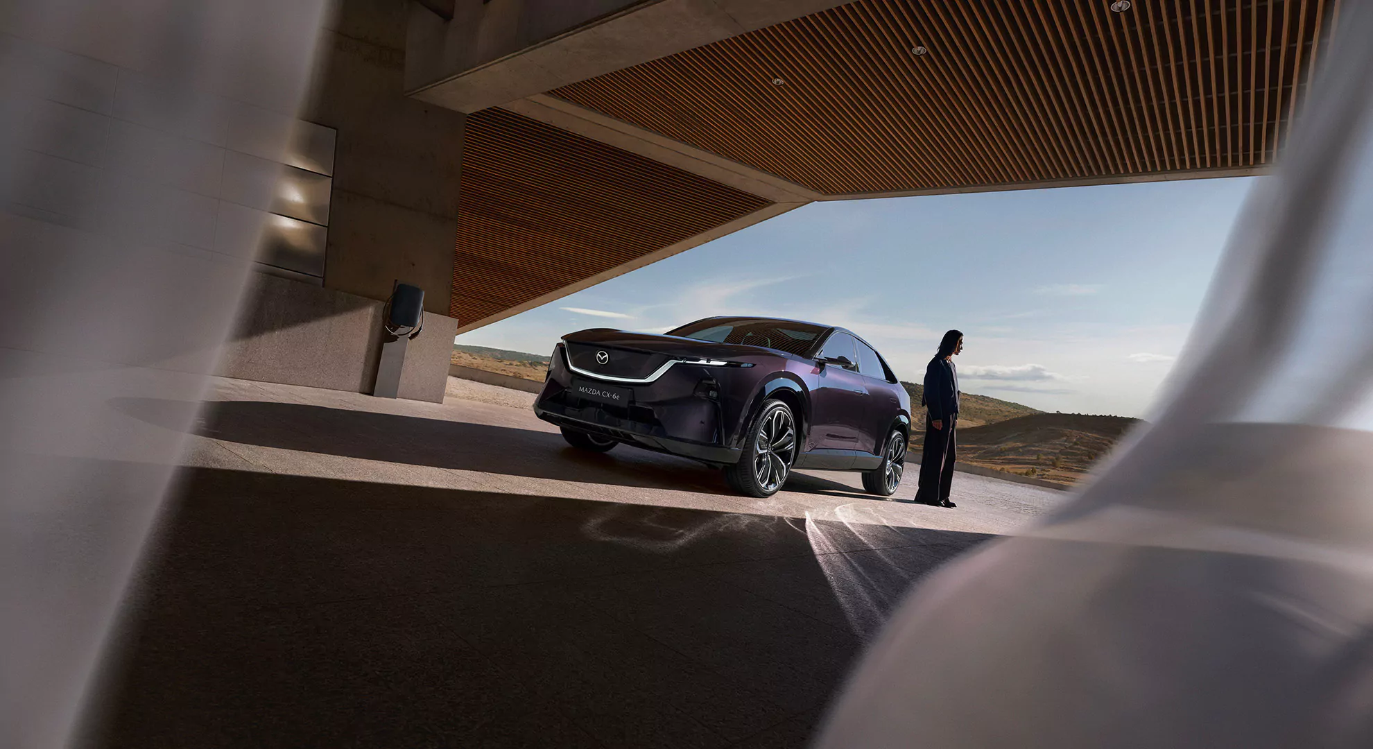 Eine Person steht neben einem dunkelvioletten Mazda MX-30-Elektroauto, das unter einem modernen Gebäude mit einer malerischen Landschaft im Hintergrund geparkt ist. Weiße Vorhänge rahmen das Bild von beiden Seiten ein.