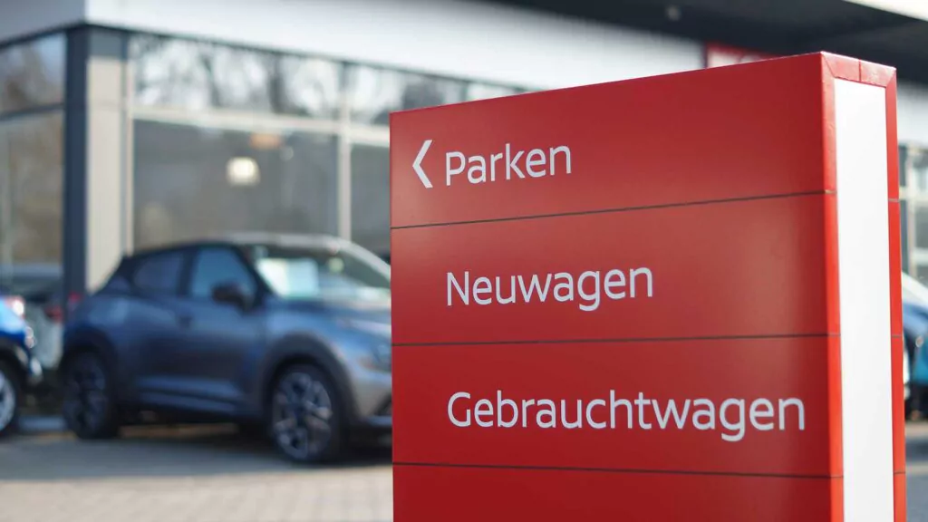 Ein rotes Schild bei einem Autohaus zeigt auf Deutsch Wegweiser zu Parkplätzen, Neuwagen und Gebrauchtwagen an; im Hintergrund sind mehrere geparkte Fahrzeuge und ein modernes Glasgebäude zu sehen.