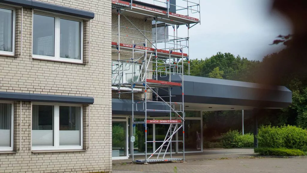 Ein Gebäude mit hellen Backsteinwänden ist teilweise von einem Metallgerüst umgeben, was auf laufende Bau- oder Renovierungsarbeiten am „Planung Gebrauchtwagen Centrum“ hindeutet. Auffällig sind die großen Fenster und der überdachte Eingang, im Hintergrund ist Grün zu sehen.