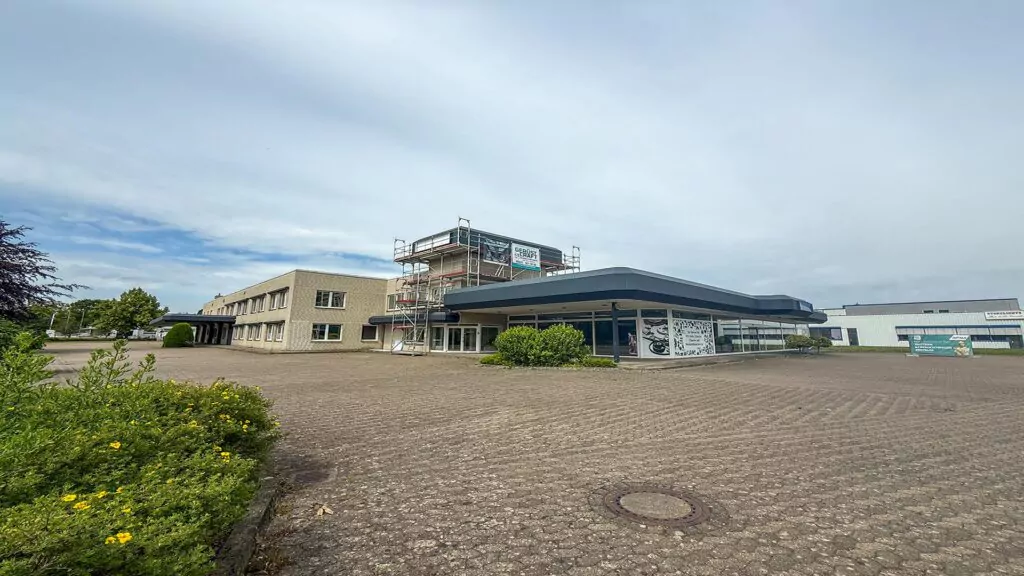 Eine große, größtenteils leere, gepflasterte Fläche vor einem modernen Gebäude mit Flachdach – wahrscheinlich Teil eines „Planung Gebrauchtwagen Centrum“ – mit einem Gerüst an einer Seite. Im Vordergrund sind etwas Grün und Büsche zu sehen, unter einem teilweise bewölkten Himmel.