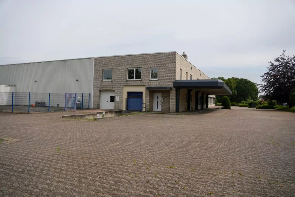 Ein großer, leerer, gepflasterter Platz vor einem beigen Industriegebäude mit blauen Akzenten lässt auf eine sorgfältige Planung des „Gebrauchtwagen-Centrums“ schließen, das über Laderampen und einen überdachten Bereich verfügt und von einem blauen Zaun sowie Grünflächen umgeben ist.