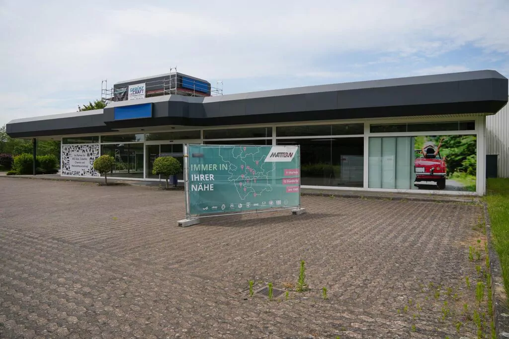 Ein modernes, leerstehendes Geschäftsgebäude mit großen Fenstern und einem Schild auf Deutsch mit der Aufschrift „IMMER IN IHRER NÄHE“. Auf dem Dach und im Inneren steht ein Gerüst, und ein Plakat mit einem roten Auto deutet auf die Planung eines künftigen Gebrauchtwagen-Centrums hin.