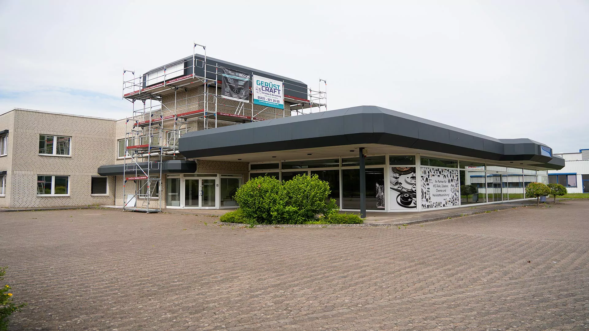 Ein modernes Gebäude, das an einer Seite mit Gerüsten versehen ist, lässt auf laufende Bauarbeiten schließen. Über den großen Fenstern befindet sich ein Schild mit der Aufschrift „Gebruikt Kantoor“; der Blick fällt auf einen größtenteils leeren, gepflasterten Platz und etwas Grünfläche in der Nähe des Eingangs – ideal für ein Gebrauchtwagenzentrum.