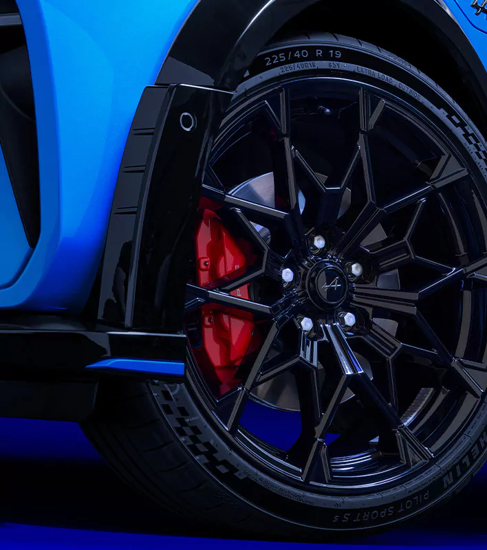 Nahaufnahme des Hinterrads eines Autos, zu sehen sind eine glänzend schwarze Mehrspeichenfelge, ein roter Bremssattel und ein Michelin Pilot Sport-Reifen an einem blauen Sportwagen, bei dem ein Teil des Kotflügels und der Seitenverkleidung zu sehen sind.