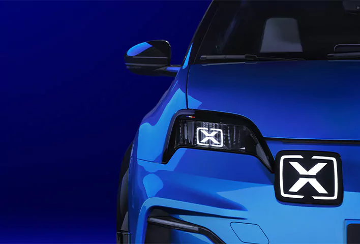 Nahaufnahme der rechten Vorderseite einer blauen Alpine A290 mit stilisierten, X-förmigen LED-Leuchten an den Scheinwerfern und am Kühlergrill vor einem einfarbig blauen Hintergrund.
