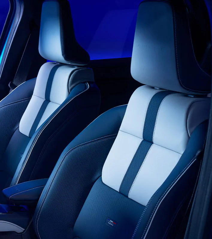 Zwei moderne Autositze mit blauer und hellgrauer Polsterung, sportlichen Ziernähten und dem in die Kopfstützen geprägten Alpine-Logo werden im Fahrzeuginneren von einer sanften blauen Beleuchtung in Szene gesetzt.