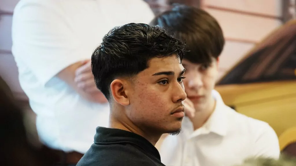 Ein junger Mann mit kurzen dunklen Haaren und Bart sitzt in einem Raum und wirkt nachdenklich, vielleicht grübelt er gerade über seine zukünftige Ausbildung 2026 nach. Im Hintergrund ist eine weitere junge Person in einem weißen Hemd zu sehen, die teilweise unscharf abgebildet ist.