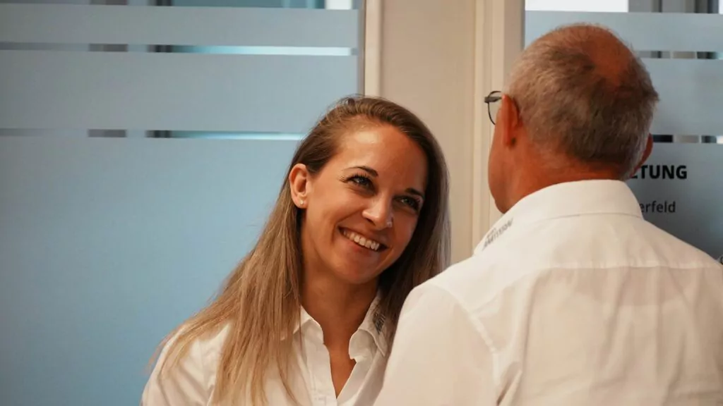 Eine Frau mit langen blonden Haaren lächelt einen Mann mit kurzen grauen Haaren herzlich an. Beide tragen weiße Hemden und stehen in einem Raum in der Nähe von Milchglasfenstern, wo sie sich freundlich über „Ausbildung 2026“ unterhalten.