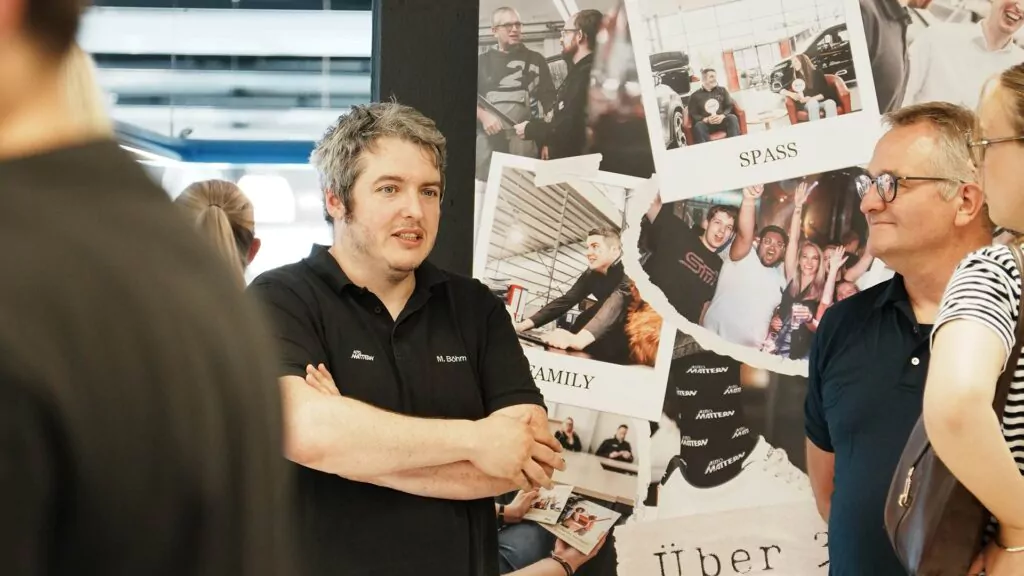 Ein Mann mit grauen Haaren und einem schwarzen Hemd steht mit verschränkten Armen da und spricht vor einer Gruppe über „Ausbildung 2026“. Hinter ihm hängt eine Fotocollage mit den Aufschriften „SPASS“ und „FAMILY“. Andere Menschen hören aufmerksam zu.