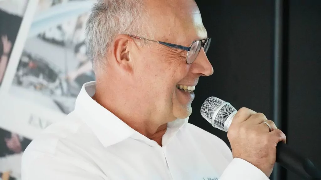 Ein lächelnder älterer Mann mit Brille und weißem Hemd spricht vor einem unscharfen Hintergrund mit verschiedenen Bildern in ein Mikrofon – möglicherweise spricht er über Chancen und Erkenntnisse im Zusammenhang mit „Ausbildung 2026“.