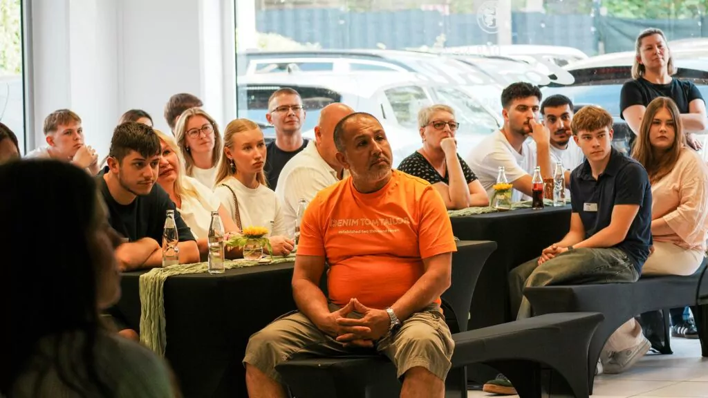 Eine Gruppe von Menschen sitzt an Tischen und hört aufmerksam jemandem zu, der nicht im Bild zu sehen ist – vielleicht diskutieren sie über Möglichkeiten wie „Ausbildung 2026“. Ein Mann in einem orangefarbenen Hemd sitzt in der Mitte. Auf den Tischen stehen Flaschen und Gläser; vor dem Fenster sind Autos zu sehen.