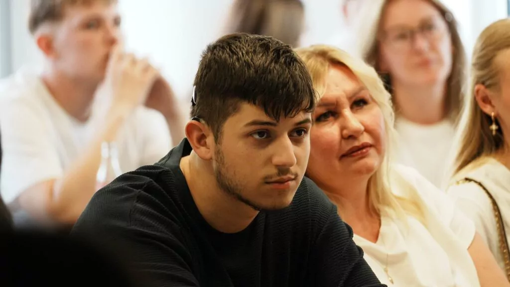 Ein junger Mann und eine ältere Frau sitzen dicht beieinander und hören aufmerksam zu, offenbar bei einer öffentlichen Veranstaltung oder einer Versammlung zum Thema „Ausbildung 2026“. Im unscharfen Hintergrund sind weitere Personen zu sehen.