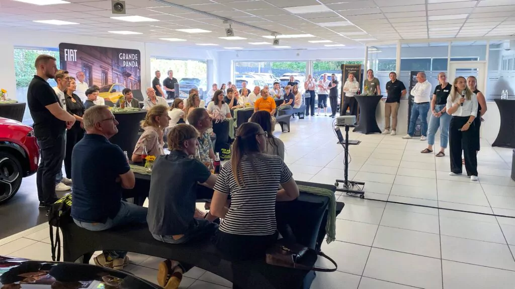 Eine Gruppe von Menschen sitzt und steht in einem hellen Ausstellungsraum und hört sich eine Präsentation zum Thema „Ausbildung 2026“ an. Zwei Frauen im Vordergrund sprechen, während die anderen zuschauen. Im Hintergrund ist eine Ausstellung mit FIAT- und GRANDE PANDA-Bannern zu sehen.