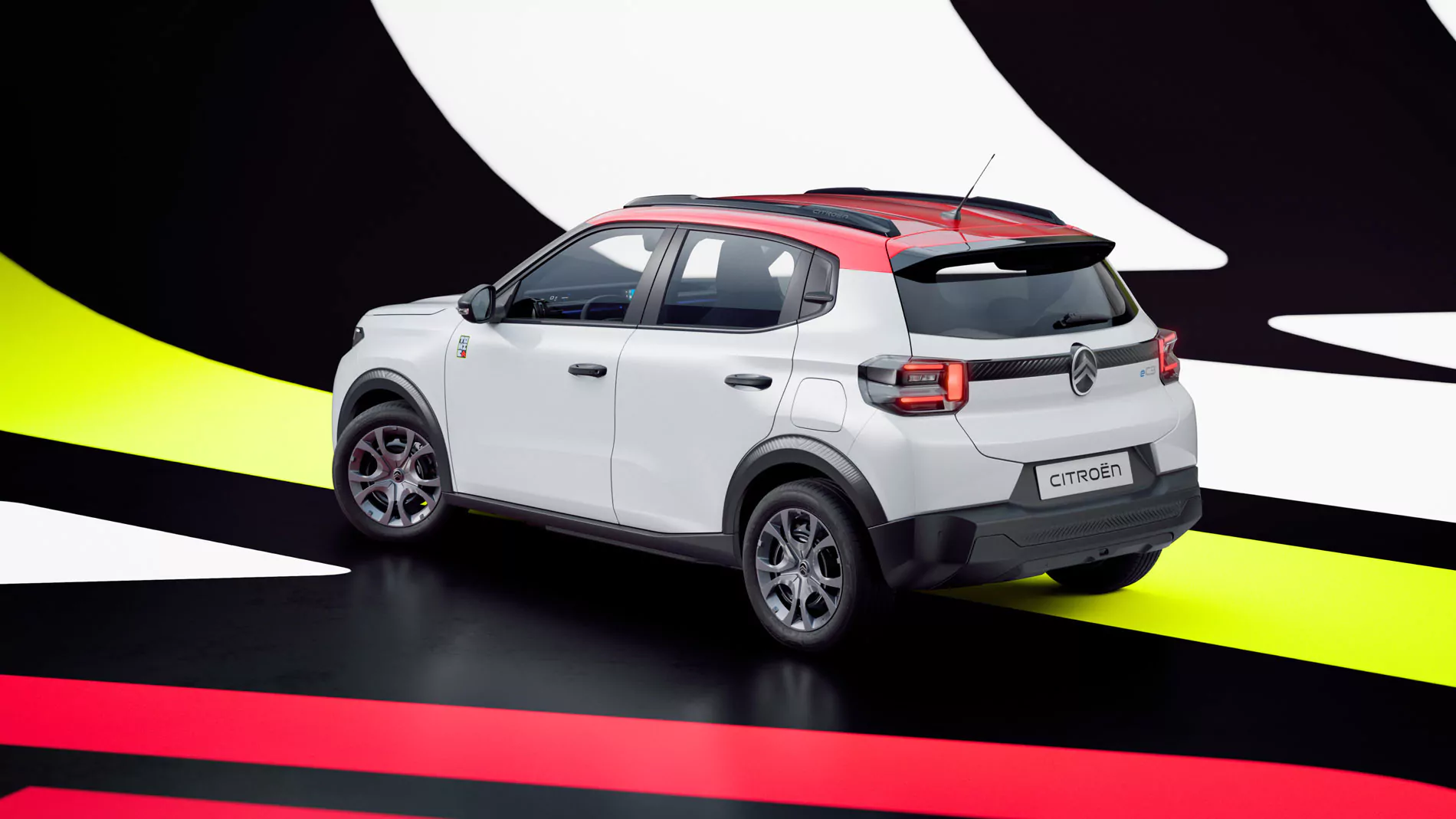 Ein weißer Citroën-SUV mit rotem Dach steht auf einem schwarzen Untergrund, im Hintergrund sind auffällige gelbe, rote und weiße grafische Muster zu sehen.