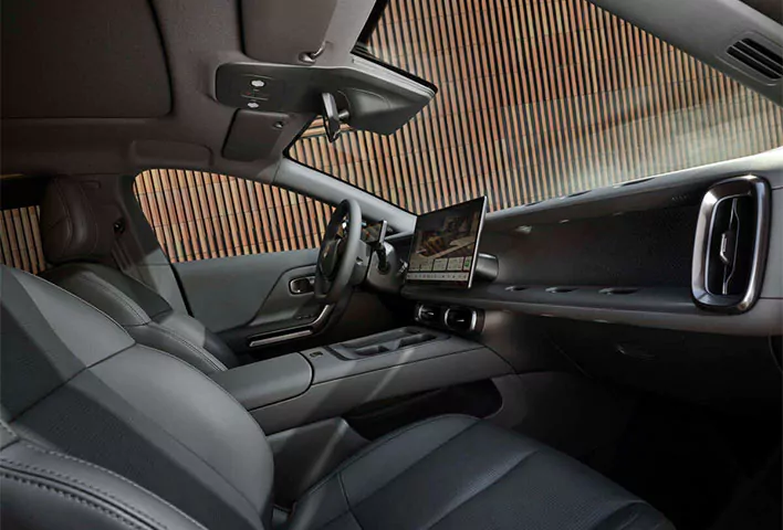 Der Innenraum eines modernen Autos wie dem Leapmotor B05 Angebot ist mit schwarzen Ledersitzen, einem breiten Armaturenbrett und einem großen Touchscreen-Display ausgestattet, das von der Beifahrerseite aus zu sehen ist. Durch die Fenster sind vertikale Holzlamellen zu erkennen.