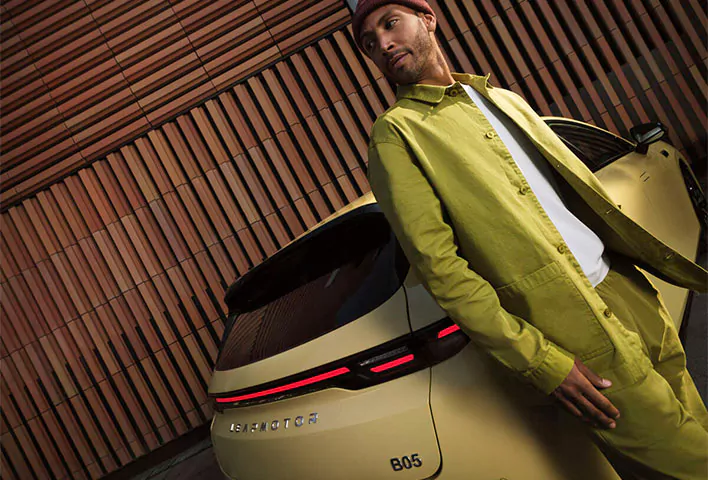 Ein Mann in grüner Kleidung lehnt an der Rückseite eines gelben Leapmotor B05 und unterstreicht damit das einzigartige Design und die moderne Ausstrahlung dieses Autos, während im Hintergrund eine elegante Wand aus Holzlamellen zu sehen ist – eine perfekte Kulisse, um das neueste Leapmotor B05-Angebot in Szene zu setzen.
