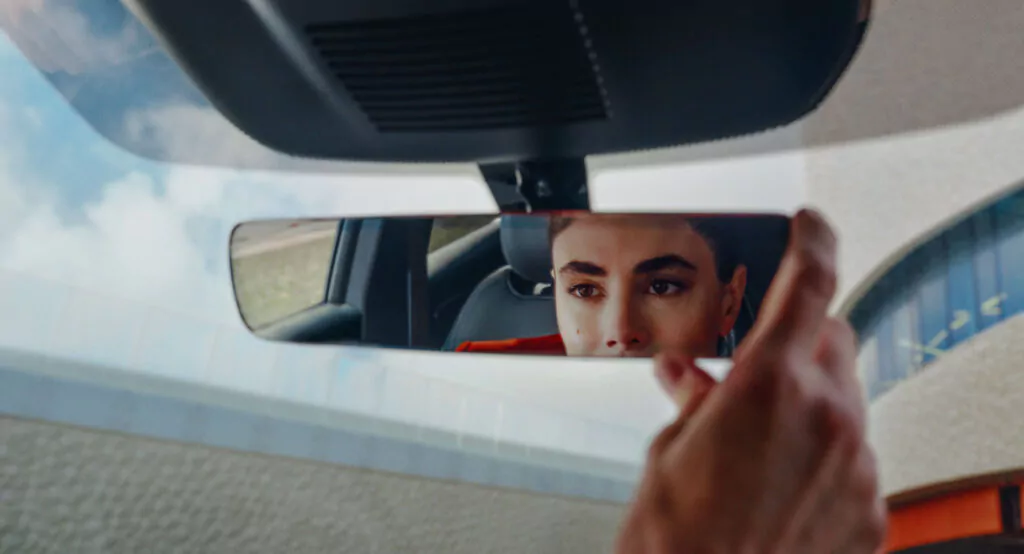 Eine Person stellt im Auto den Rückspiegel ein, in dem sich ihr konzentrierter Gesichtsausdruck widerspiegelt, während sie nach vorne blickt. Durch die Fenster sind der Dachhimmel des Autos und ein Gebäude im Freien zu sehen.